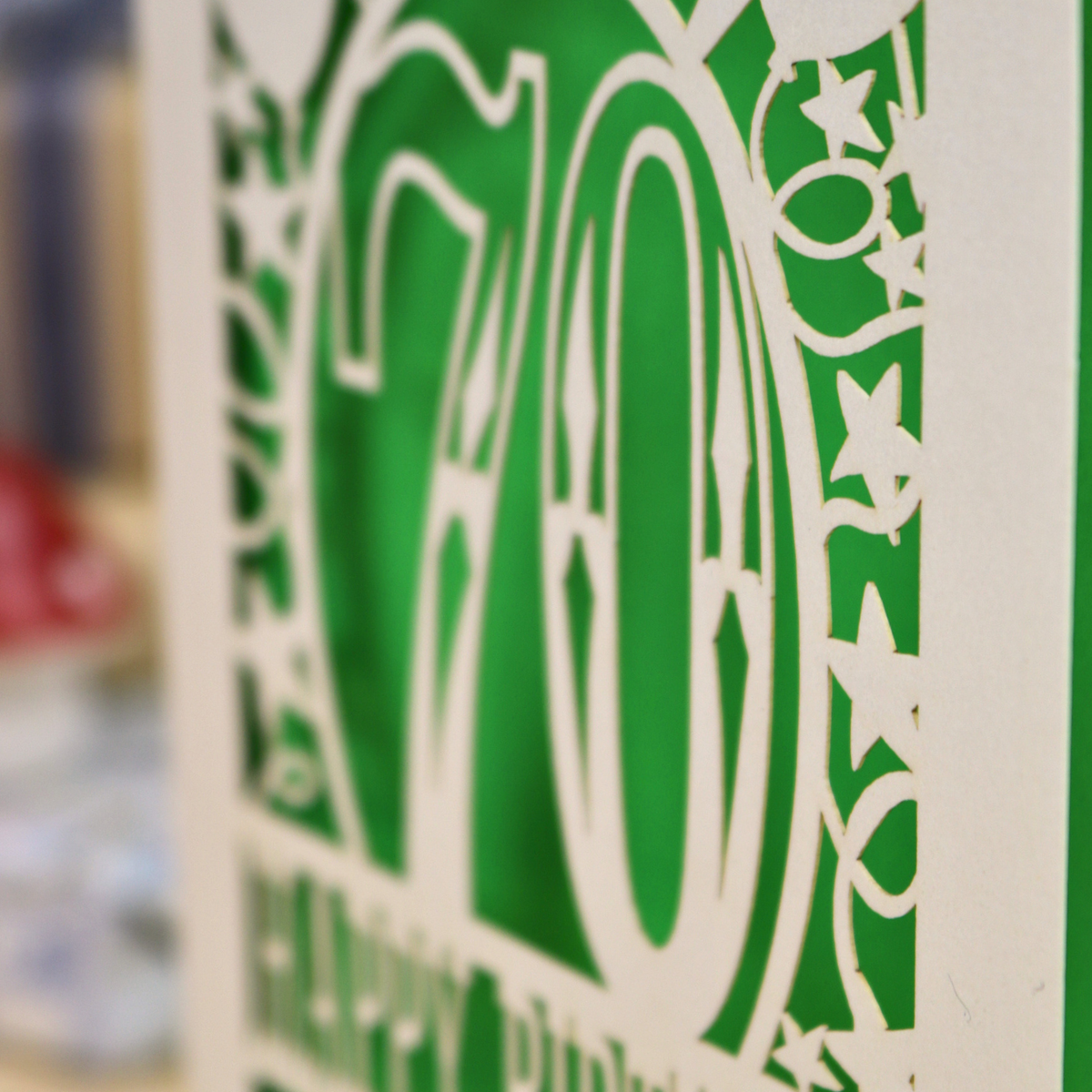 A close-up of a green and cream decorative birthday card featuring the number 70 and cut out stars and scrollwork, with blurred background elements.