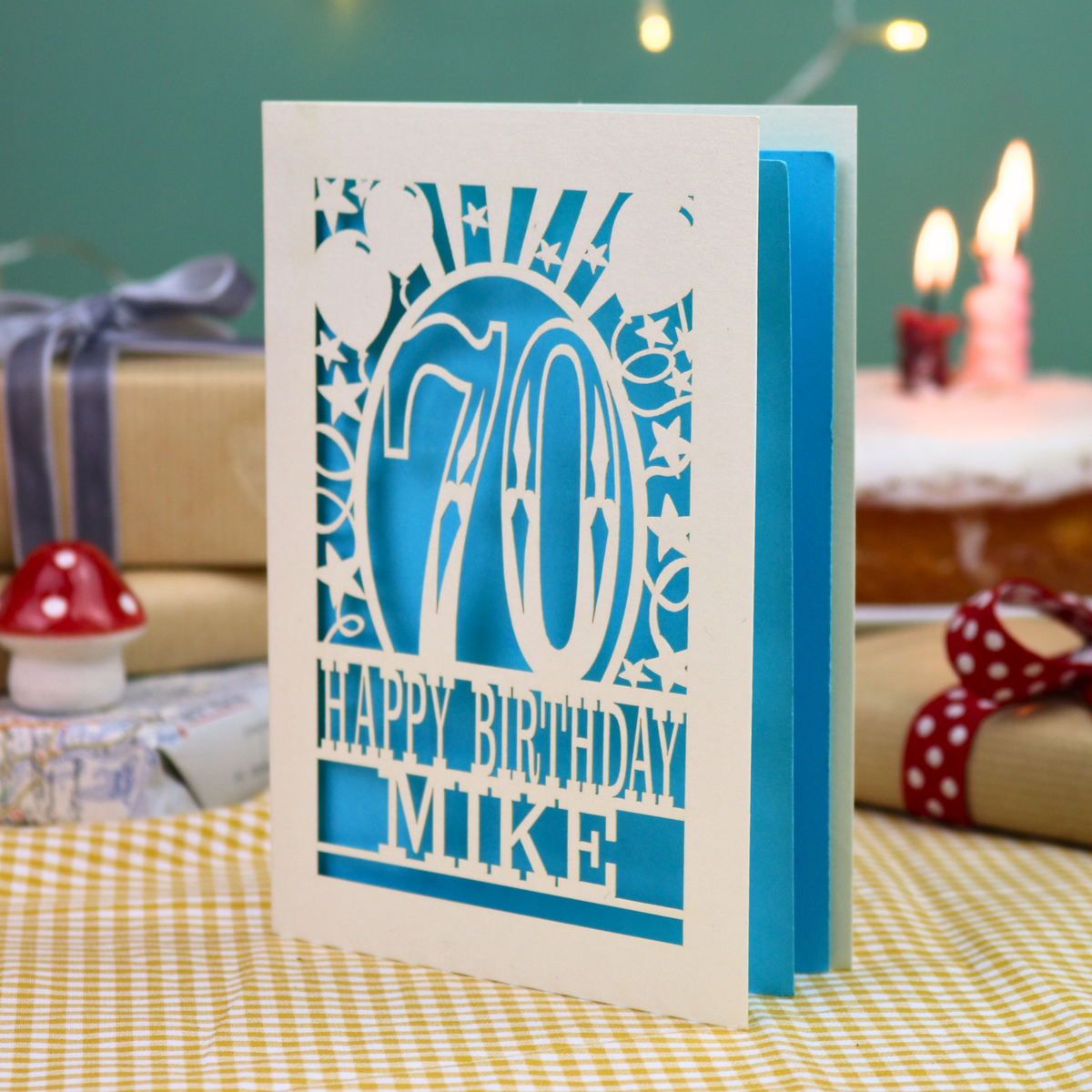 A personalised laser-cut 70th birthday card, featuring a cream cut out '70' design with blue backing and the text 'HAPPY BIRTHDAY MIKE', displayed on a checked tablecloth with blurred birthday decorations in the background.