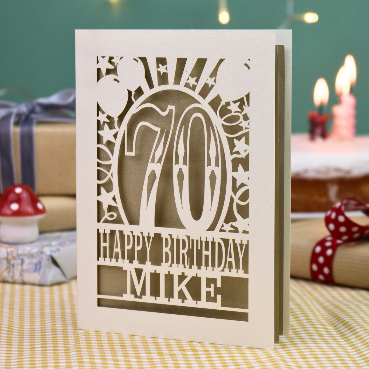 A personalised laser-cut birthday card for a 70th birthday, featuring a cream cut out '70' design with gold leaf backing and the text 'HAPPY BIRTHDAY MIKE', displayed on a checked tablecloth with blurred birthday decorations in the background.