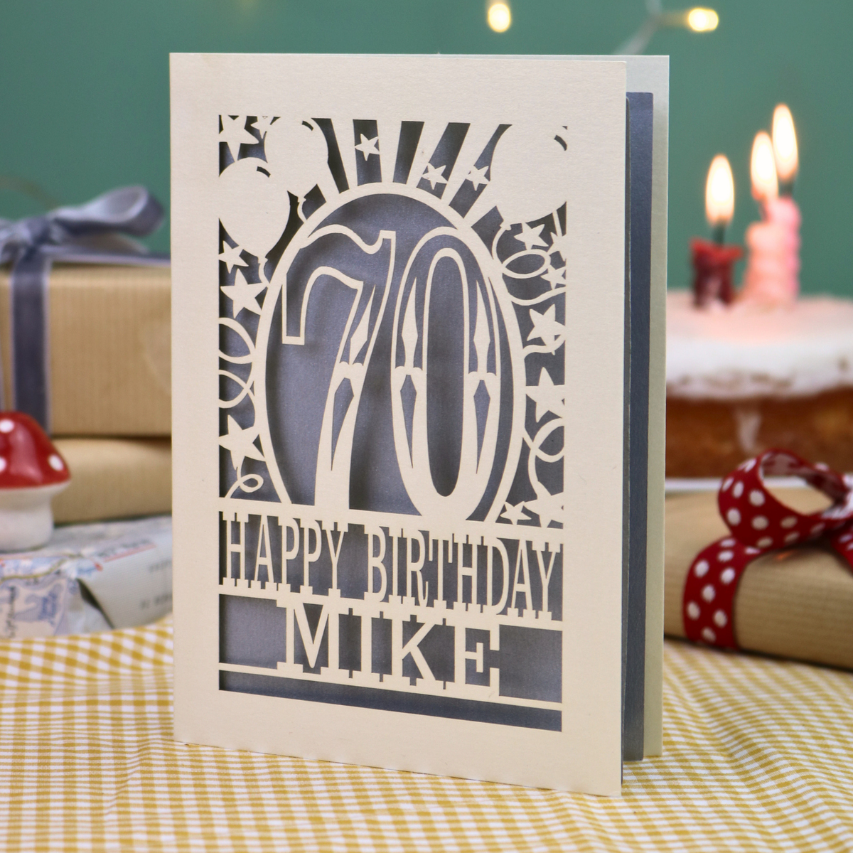 A laser-cut 70th birthday card standing upright on a checked tablecloth, surrounded by gift boxes and lit candles in the background.