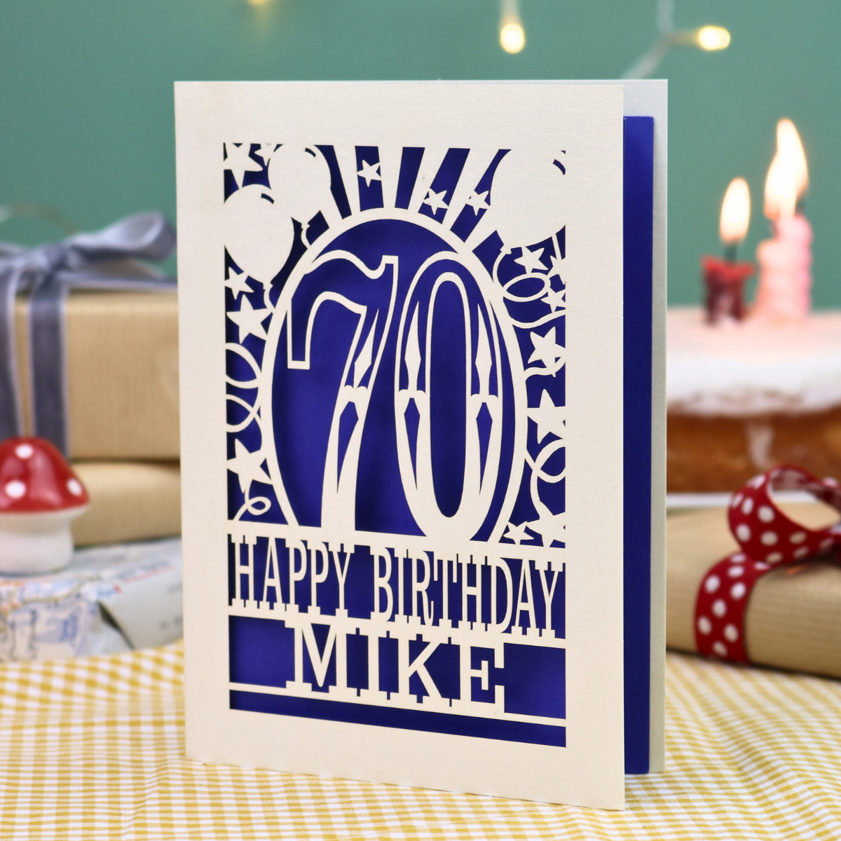 A personalized 70th birthday laser-cut greeting card for 'Mike' standing upright on a yellow-and-white checked tablecloth. The card features a cream card with intricate violet laser-cut designs, including stars, stripes, balloons, and the number 70.