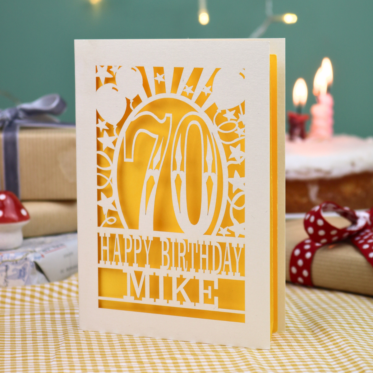 A personalized laser-cut birthday card for a 70th birthday, featuring a cream card with yellow paper and laser-cut out details spelling '70' and 'HAPPY BIRTHDAY MIKE'. The card is displayed on a yellow-and-white checked tablecloth with a blurred birthday cake and candles in the background.