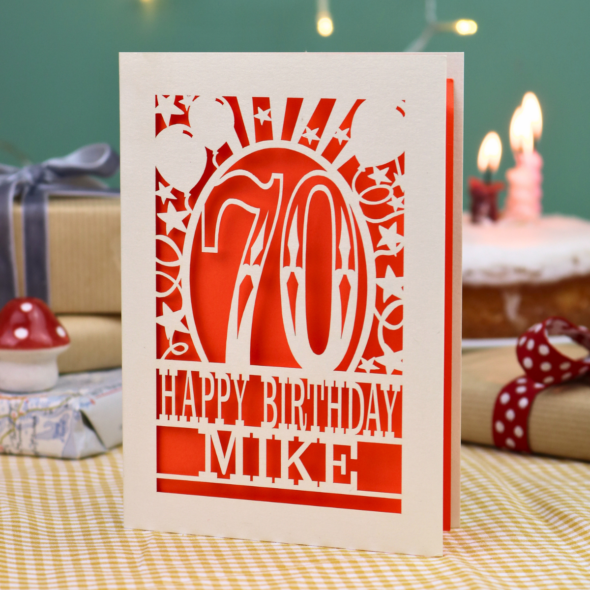 A personalised laser-cut 70th birthday card, featuring a cream cut out '70' design with orange backing and the text 'HAPPY BIRTHDAY MIKE', displayed on a checked tablecloth with blurred birthday decorations in the background.