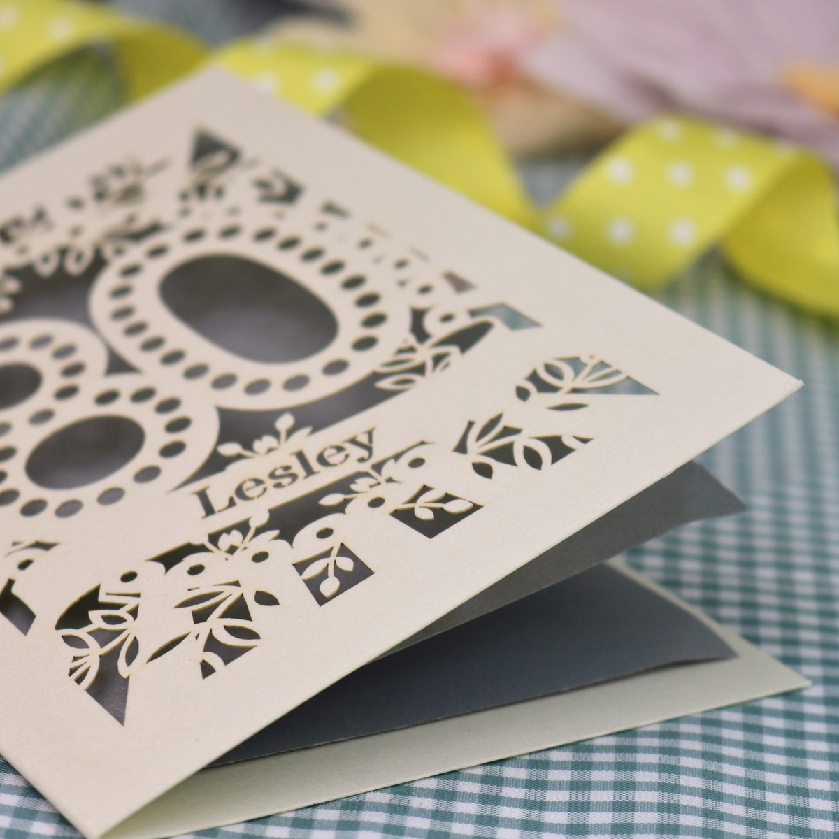 A personalized laser-cut 80th birthday card  featuring intricate cream cut out floral patterns and the number '80' with the name 'Lesley' on a banner