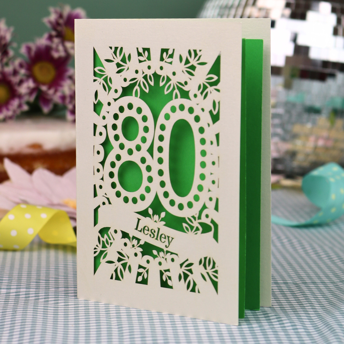 A personalized laser-cut 80th birthday card  featuring intricate cream cut out floral patterns and the number '80' with the name 'Lesley' on a banner, displayed on a checked tablecloth with blurred decorative flowers in the background.