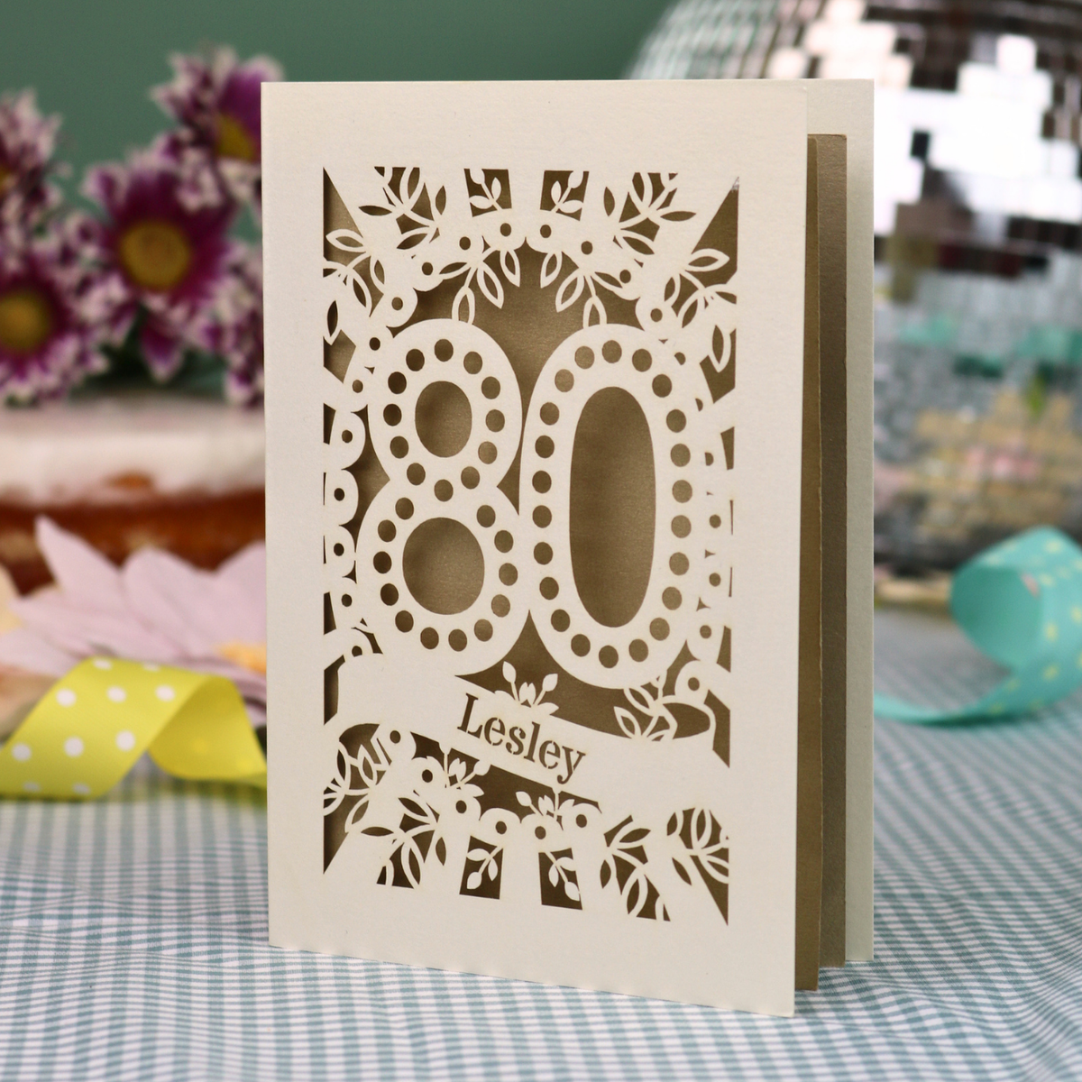A personalized laser-cut 80th birthday card  featuring intricate cream cut out floral patterns and the number '80' with the name 'Lesley' on a banner, displayed on a checked tablecloth with blurred decorative flowers in the background.