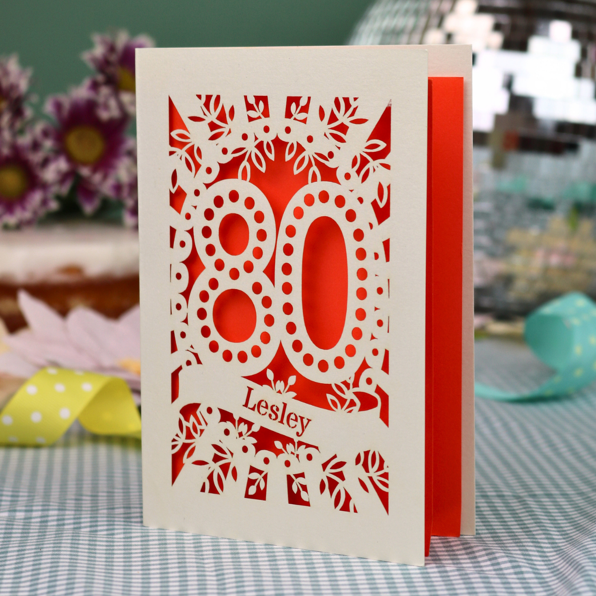 A personalized laser-cut 80th birthday card  featuring intricate cream cut out floral patterns and the number '80' with the name 'Lesley' on a banner, displayed on a checked tablecloth with blurred decorative flowers in the background.