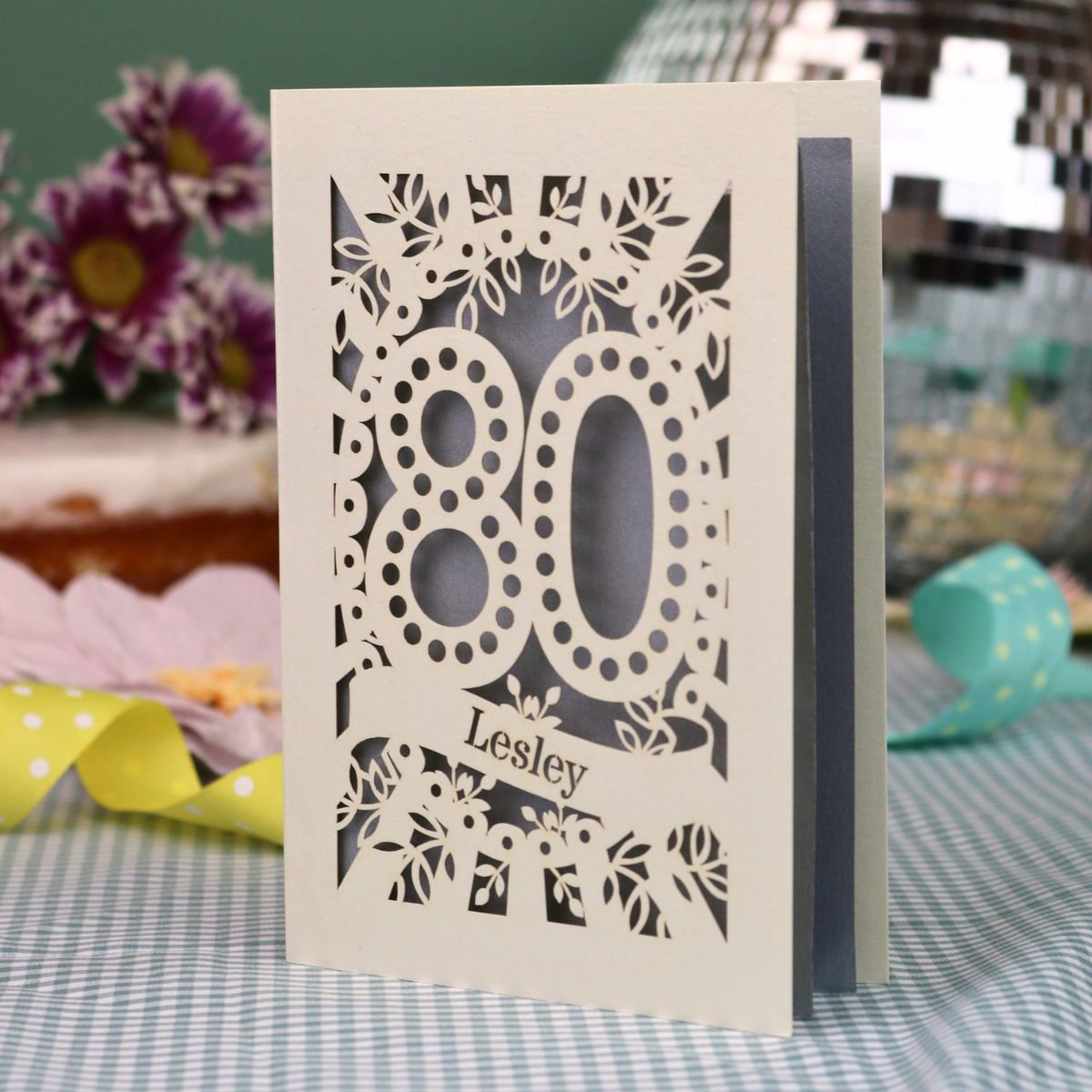 A personalized laser-cut 80th birthday card  featuring intricate cream cut out floral patterns and the number '80' with the name 'Lesley' on a banner, displayed on a checked tablecloth with blurred decorative flowers in the background.