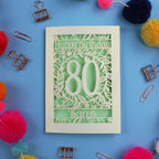 Laser Cut Birthday card with 'Happy Birthday 80 Karen' on a blue background with colourful pom-poms and paper clips.