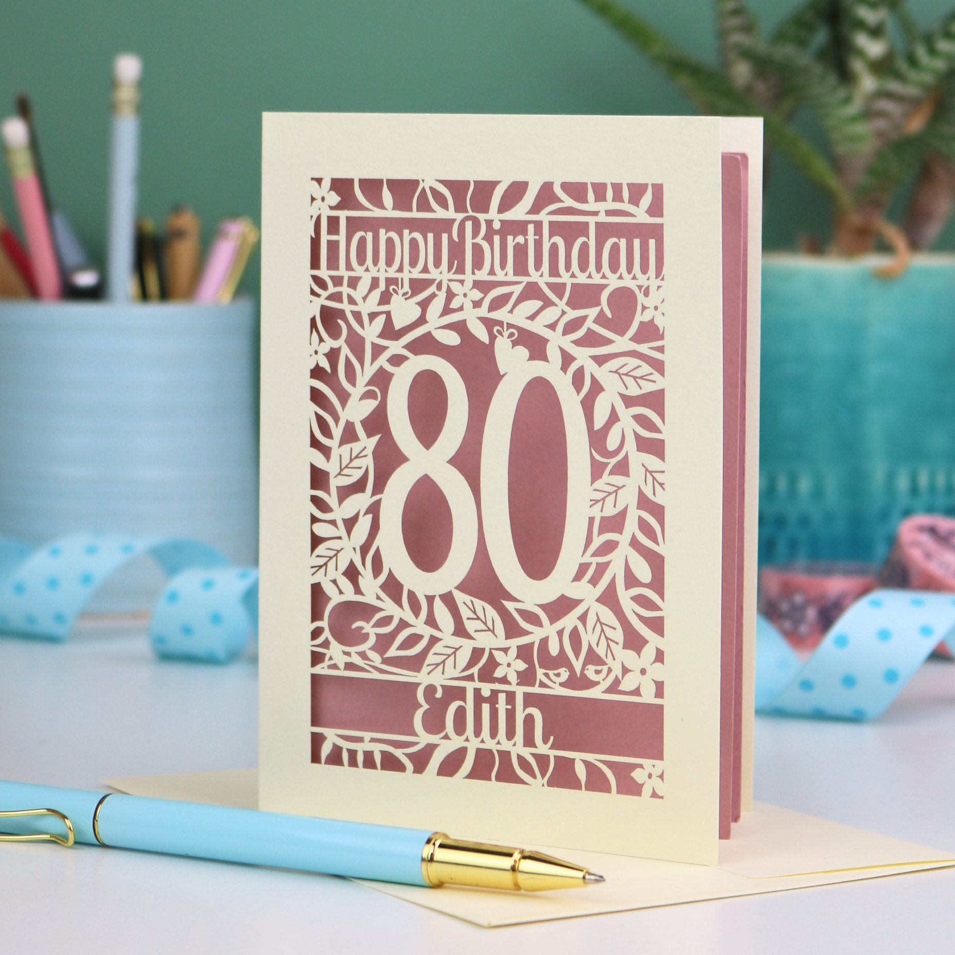 Paper-cut birthday card for 80th birthday with decorative elements on a desk. Card shows a decorative number '80' surrounded by a leafy border and 'Happy Birthday' above, with 'Edith' below