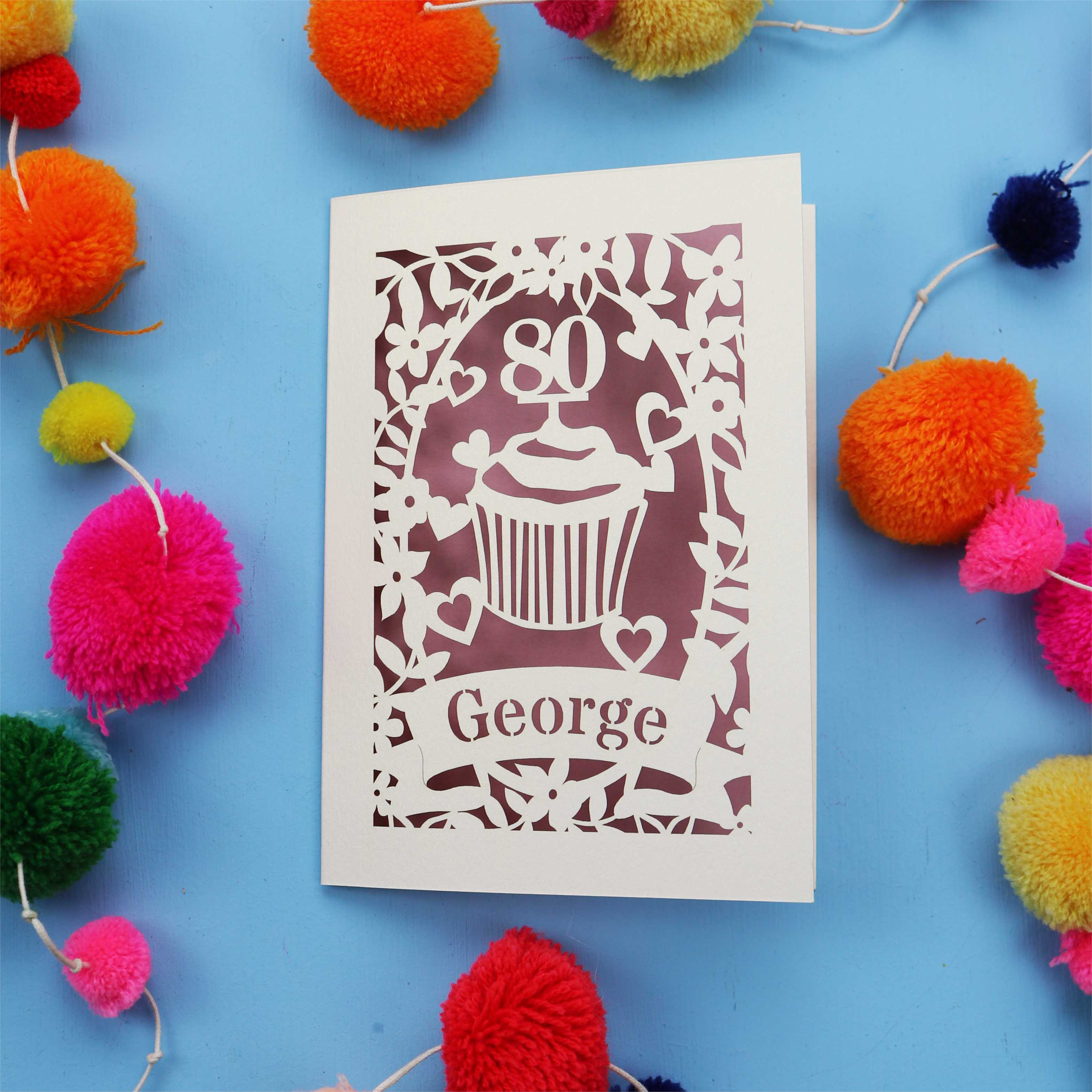 Paper cut 80th Birthday Card with '80' cut out cupcake design on a blue background with colourful pom-poms.