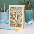 Laser-cut 80th birthday card with decorative elements on a desk. Card shows a decorative number '80' surrounded by a leafy border and 'Happy Birthday' above, with 'Edith' below