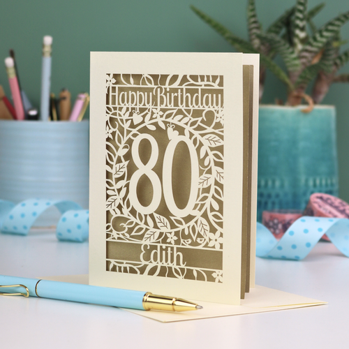 Laser-cut 80th birthday card with decorative elements on a desk. Card shows a decorative number '80' surrounded by a leafy border and 'Happy Birthday' above, with 'Edith' below