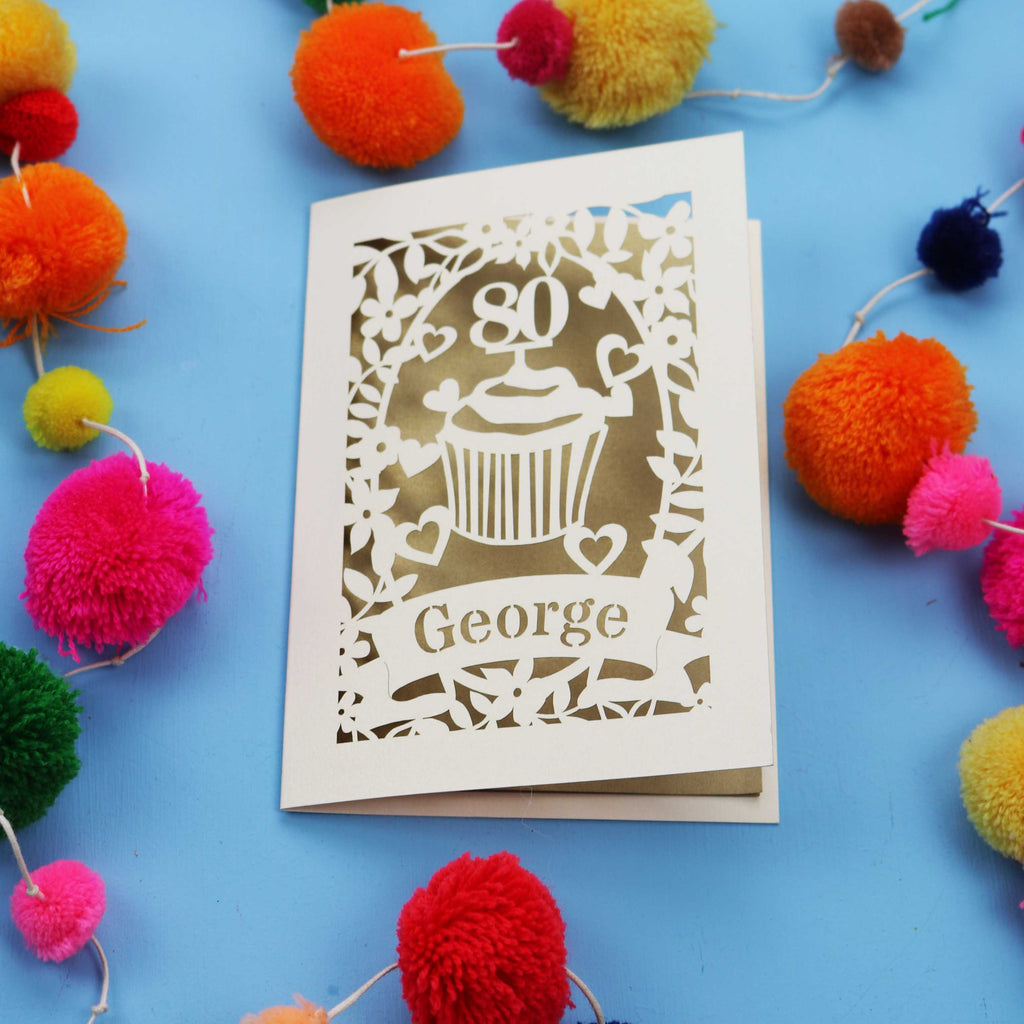 Laser cut 80th Birthday Card with '80' cut out cupcake design on a blue background with colourful pom-poms.