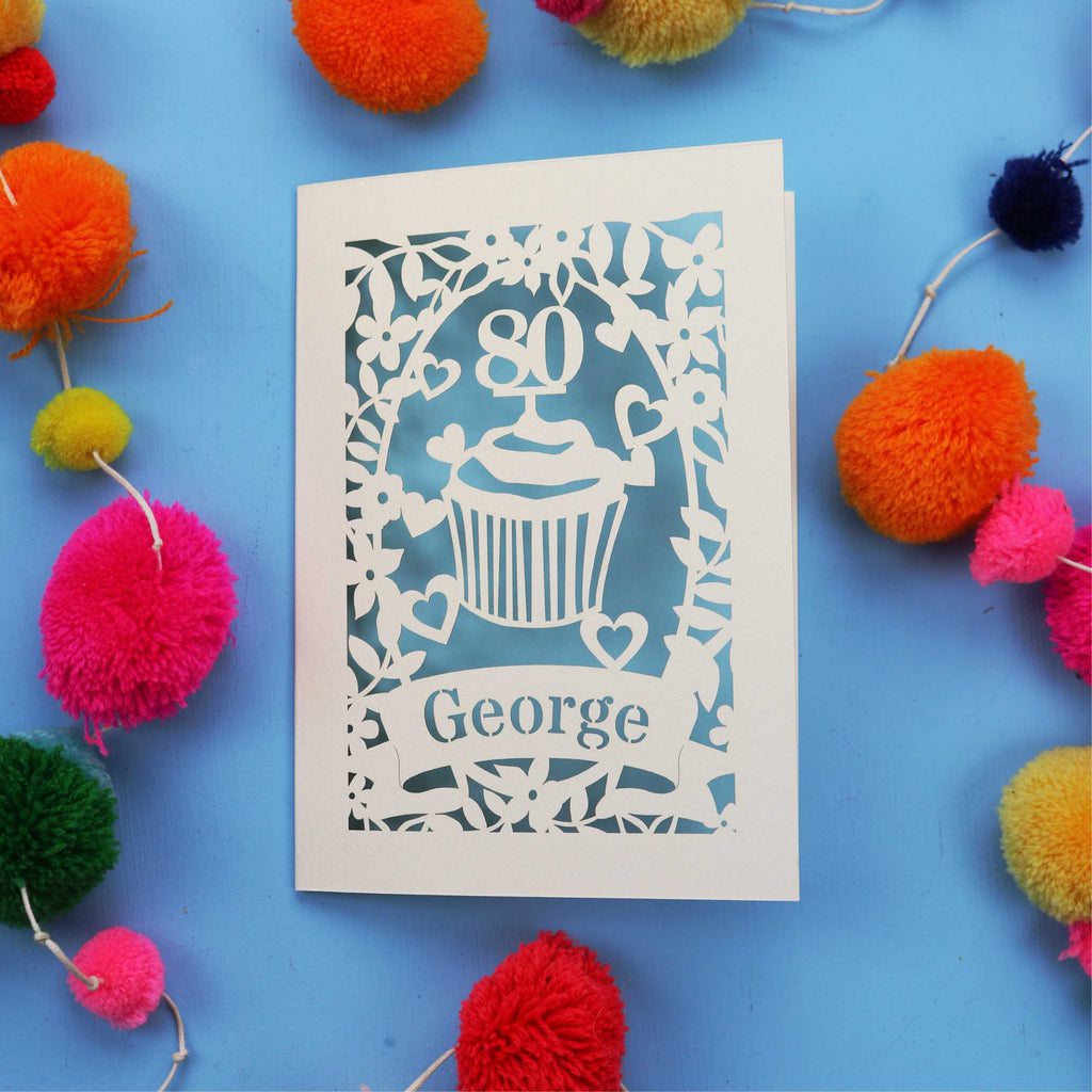 Laser cut 80th Birthday Card with '80' cut out cupcake design on a blue background with colourful pom-poms.