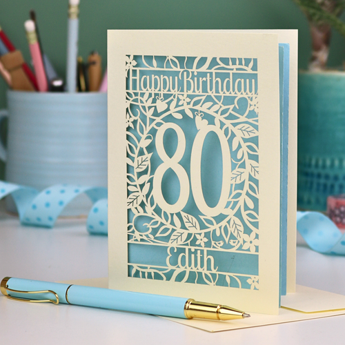 Laser-cut birthday card for 80th birthday with decorative elements on a desk. Card shows a decorative number '80' surrounded by a leafy border and 'Happy Birthday' above, with 'Edith' below