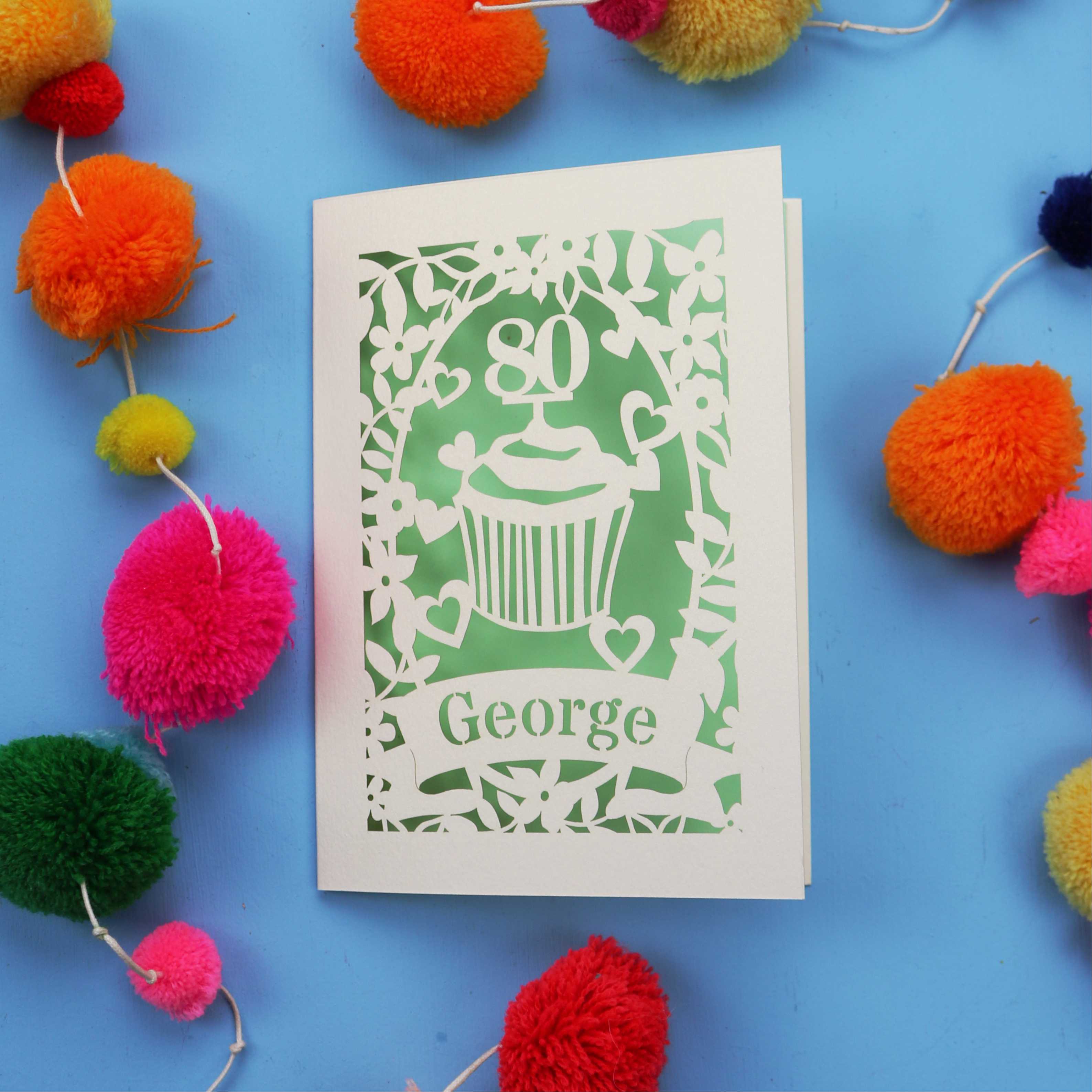 Laser cut Birthday Card with '80' cut out cupcake design on a blue background with colourful pom-poms.