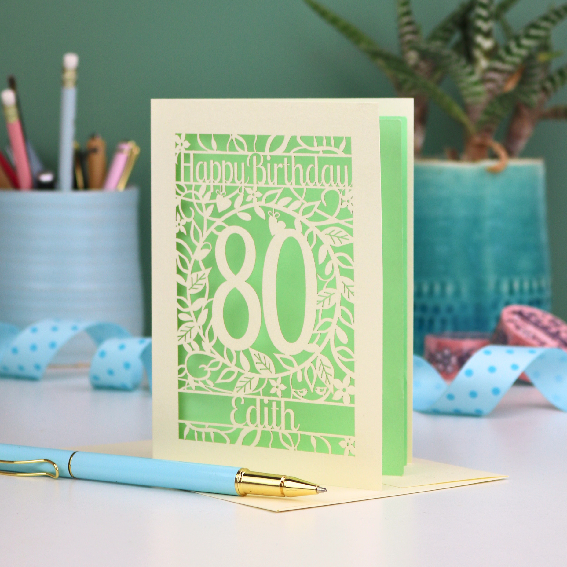 Laser-cut birthday card for 80th birthday with decorative elements on a desk. Card shows a decorative number '80' surrounded by a leafy border and 'Happy Birthday' above, with 'Edith' below