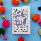 Laser cut 80th Birthday Card with '80' cut out cupcake design on a blue background with colourful pom-poms.