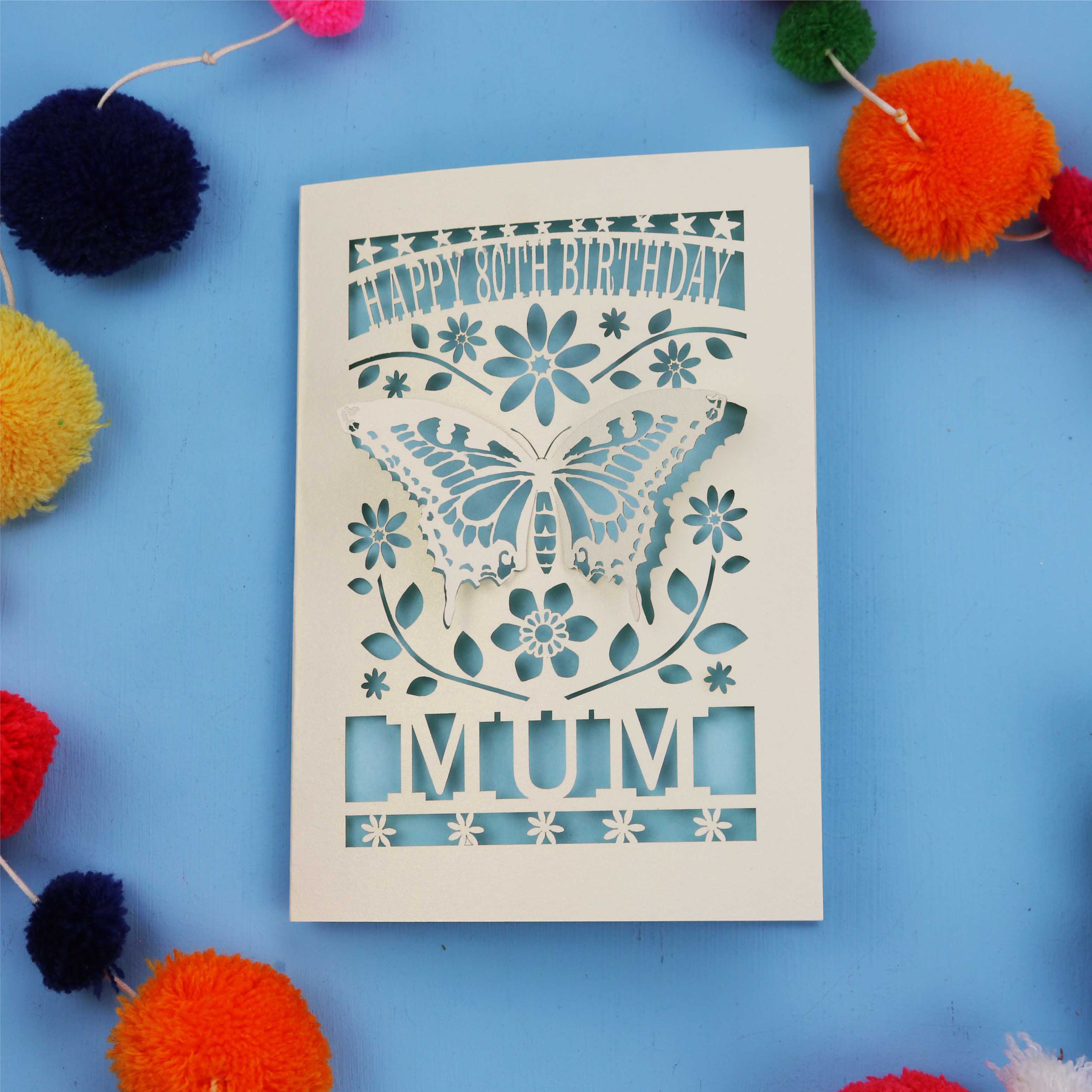 80th Birthday Card with butterfly design and 'Happy Birthday Mum' text on a blue background with colourful pom-poms.