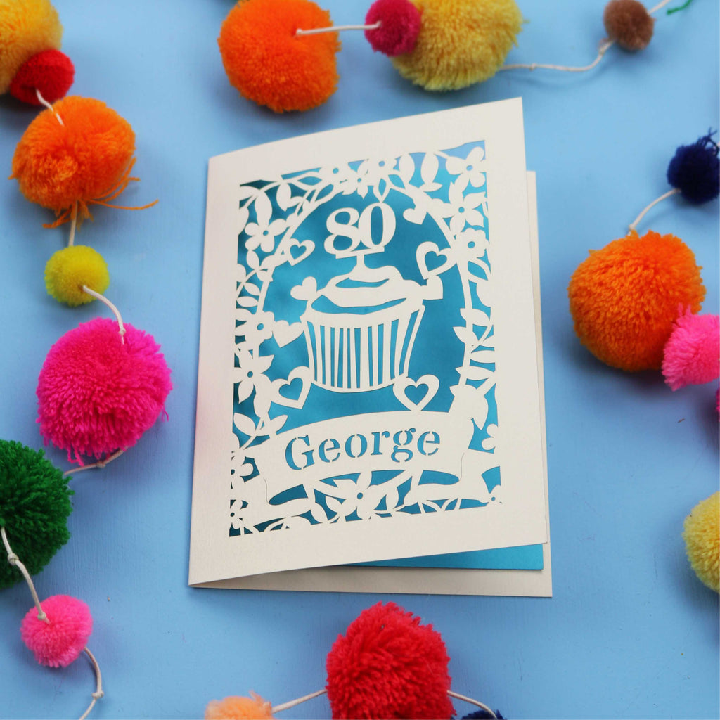Personalised Laser cut 80th Birthday Card with '80' cut out cupcake design on a blue background with colourful pom-poms.