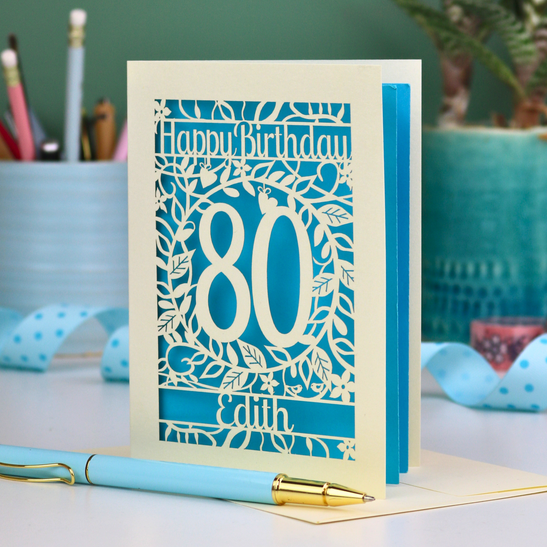 Personalised cut out birthday card for 80th birthday with decorative elements on a desk. Card shows a decorative number '80' surrounded by a leafy border and 'Happy Birthday' above, with 'Edith' below