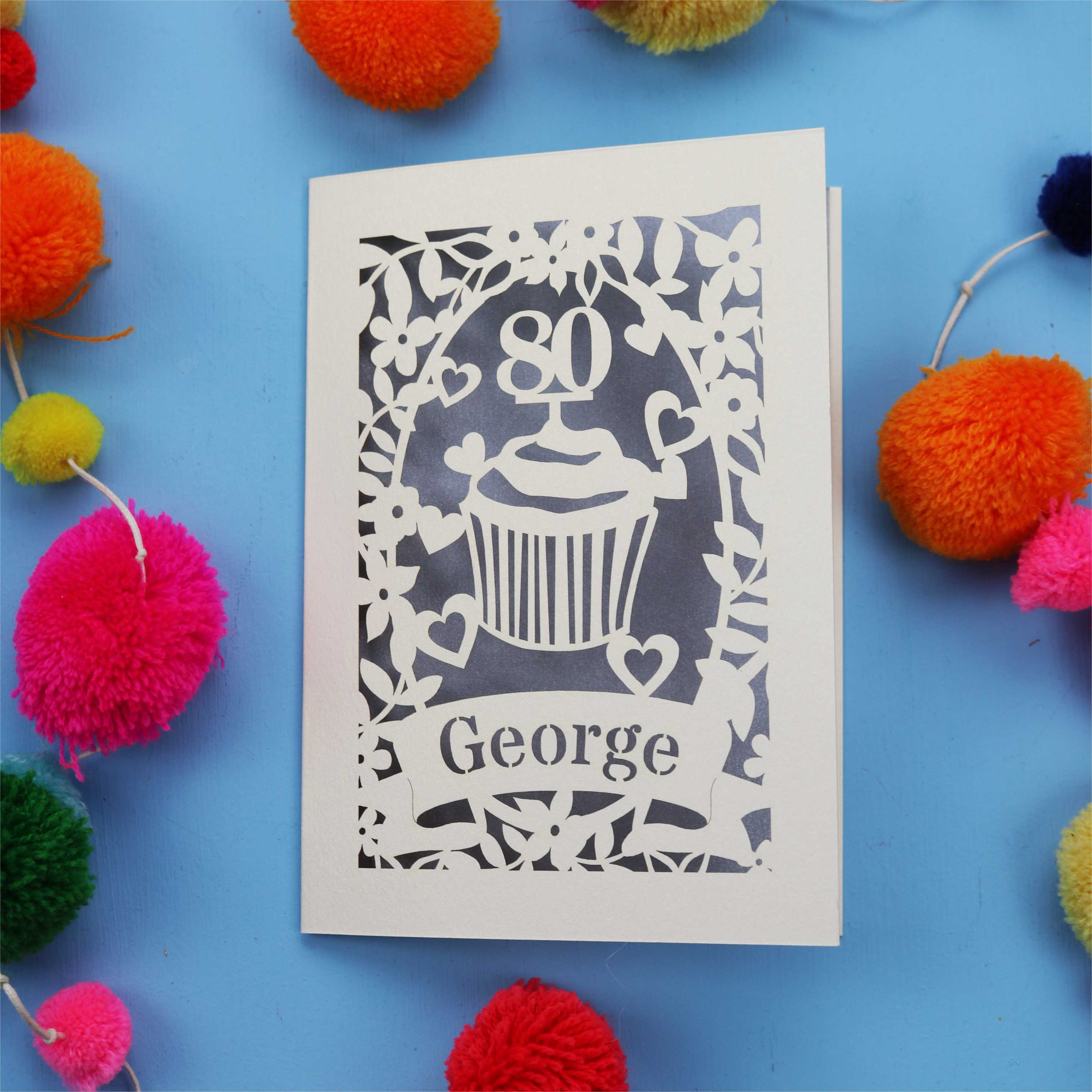 Laser cut personalised 80th Birthday Card with '80' cut out cupcake design on a blue background with colourful pom-poms.
