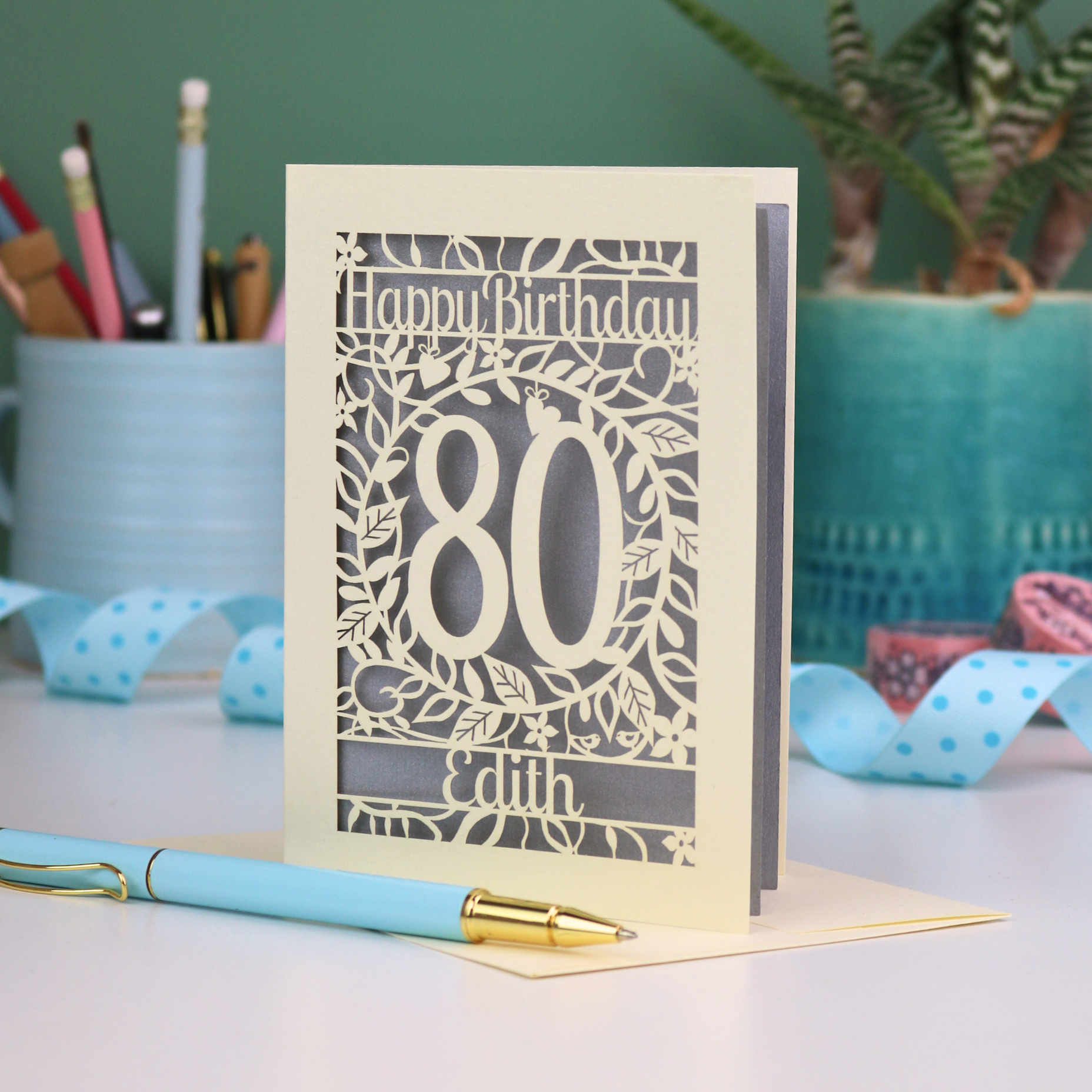 Laser-cut birthday card for 80th birthday with decorative elements on a desk. Card shows a decorative number '80' surrounded by a leafy border and 'Happy Birthday' above, with 'Edith' below