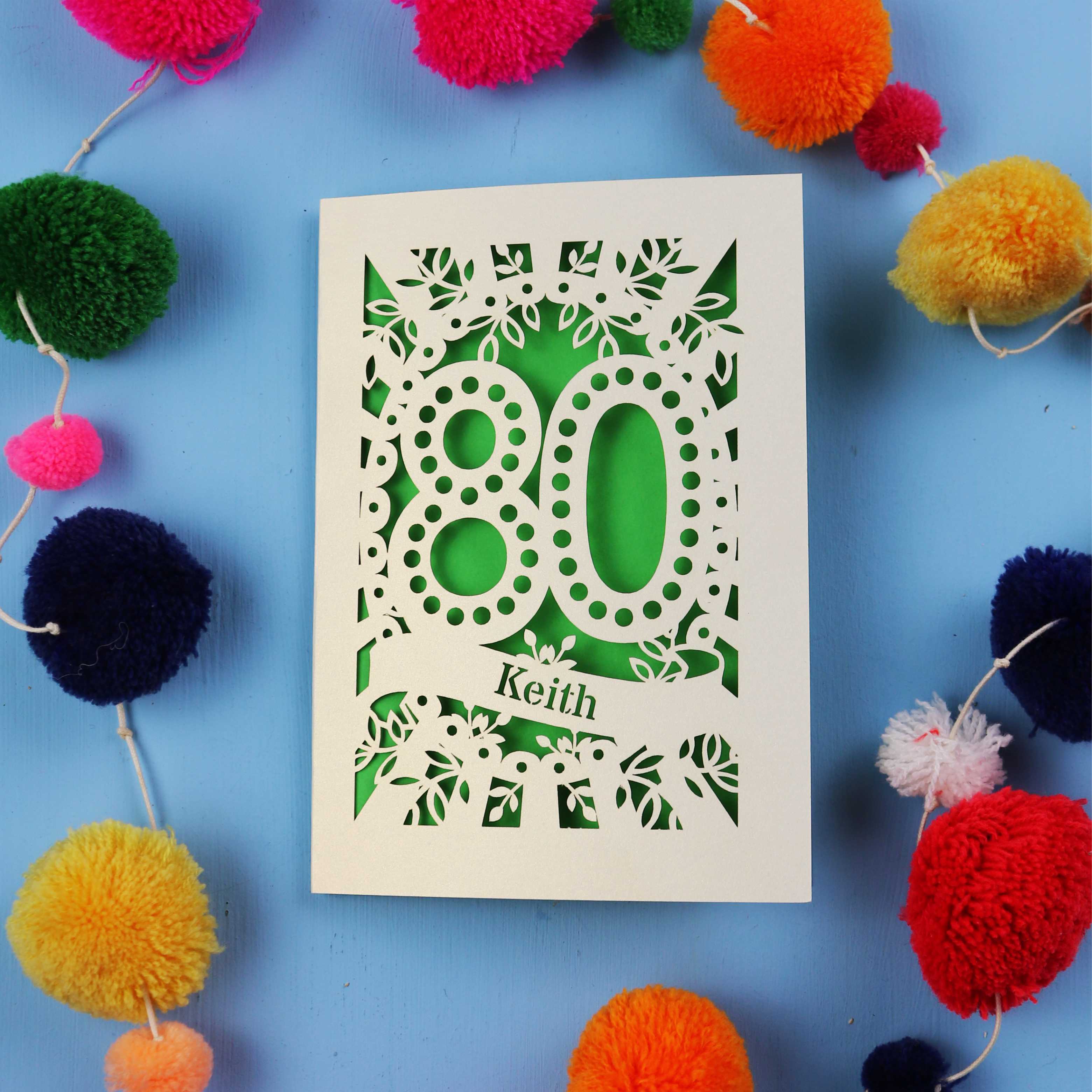 Laser cut 80th Birthday Card with '80' cut out design on a blue background with colourful pom-poms.