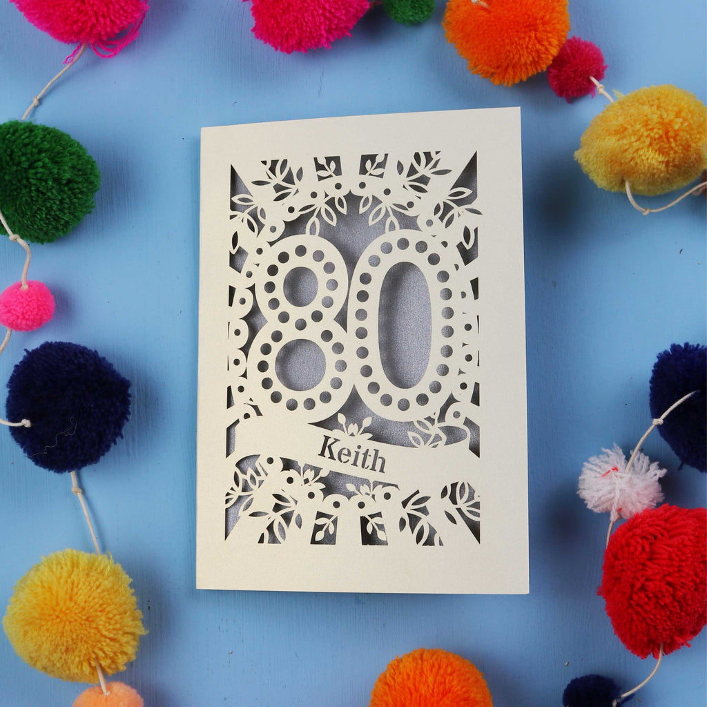 Laser cut 80th Birthday Card with '80' cut out design on a blue background with colourful pom-poms.