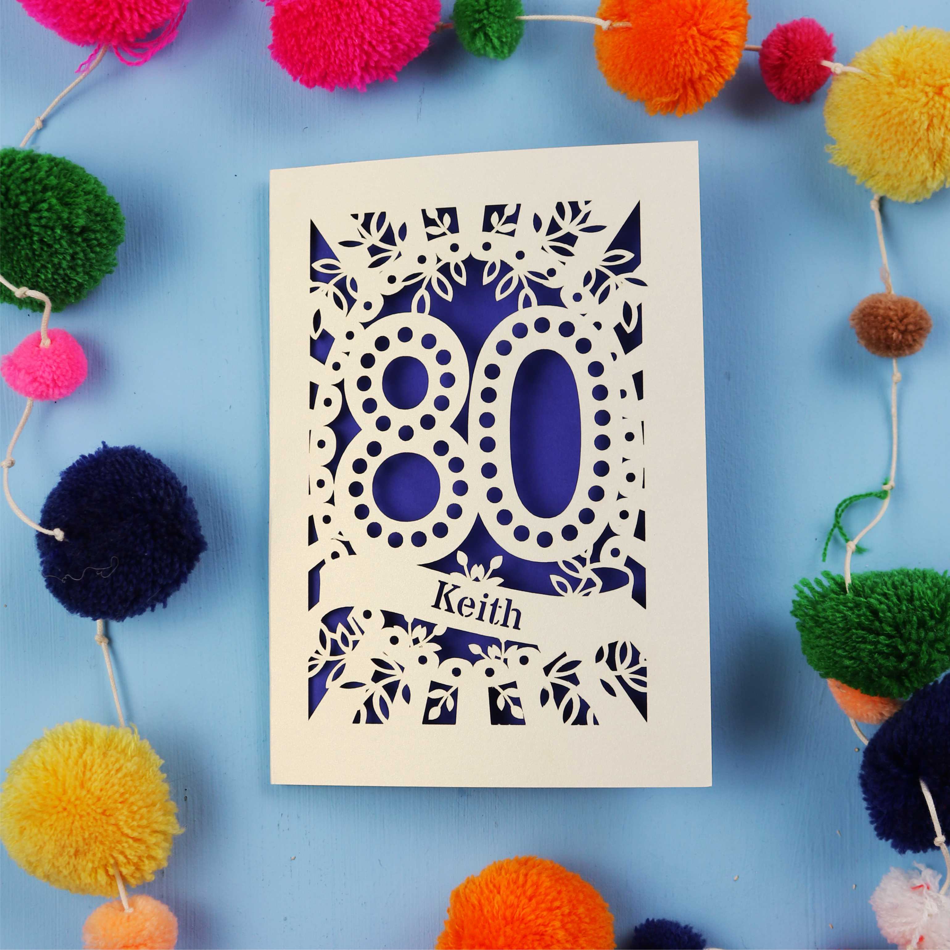Laser cut 80th Birthday Card with '80' cut out design on a blue background with colourful pom-poms.