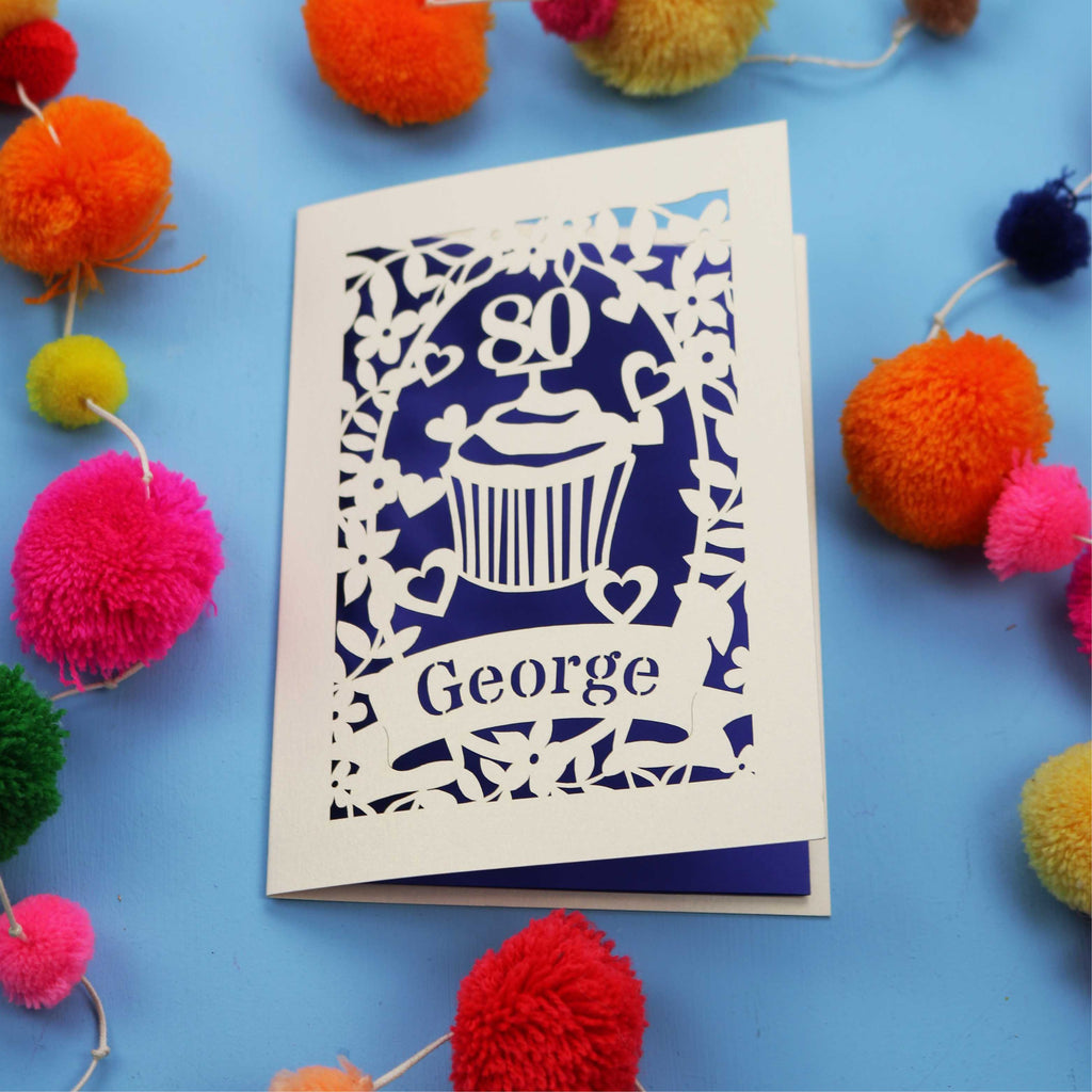Laser cut 80th Birthday Card with '80' cut out cupcake design on a blue background with colourful pom-poms.