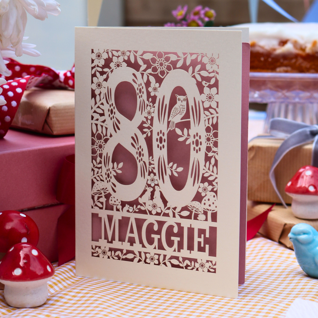 A personalized laser-cut 80th birthday card with floral and bird motifs, standing on a checkered tablecloth with gift boxes and decorative mushrooms in the background.