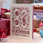 A personalized laser-cut 80th birthday card with floral and bird motifs, standing on a checkered tablecloth with gift boxes and decorative mushrooms in the background.
