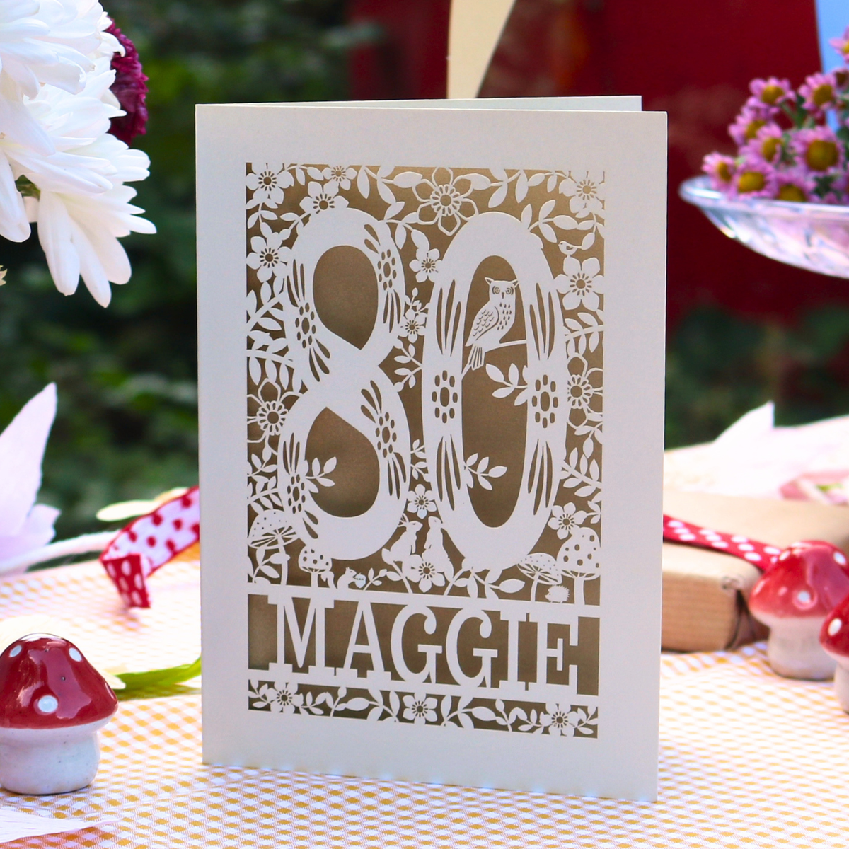 A white greeting card with laser-cut floral details and the text '80 Maggie' in the center, displayed on a checkered tablecloth with decorative flowers and small mushroom figurines in a garden setting.