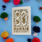 Laser cut 80th Birthday card with '80 Jean' design on a blue background with colourful pom-poms.