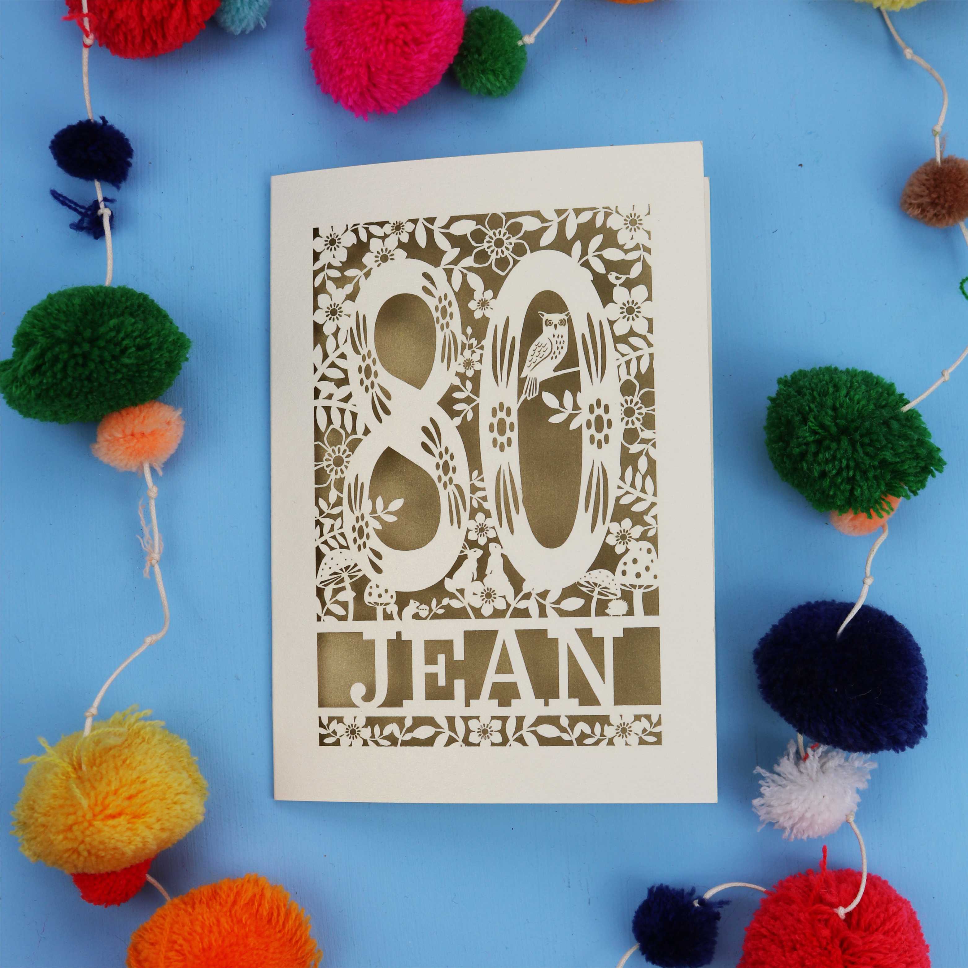 Laser cut 80th Birthday card with '80 Jean' design on a blue background with colourful pom-poms.