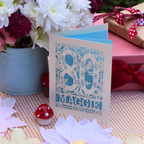 A laser cut greeting card with a blue floral pattern and the text '80th Maggie' stands on a checked tablecloth. The card is styled with paper flowers, a small red mushroom figurine, a pink gift box with a red polka-dot ribbon, and a vase of white and burgundy flowers in the background.