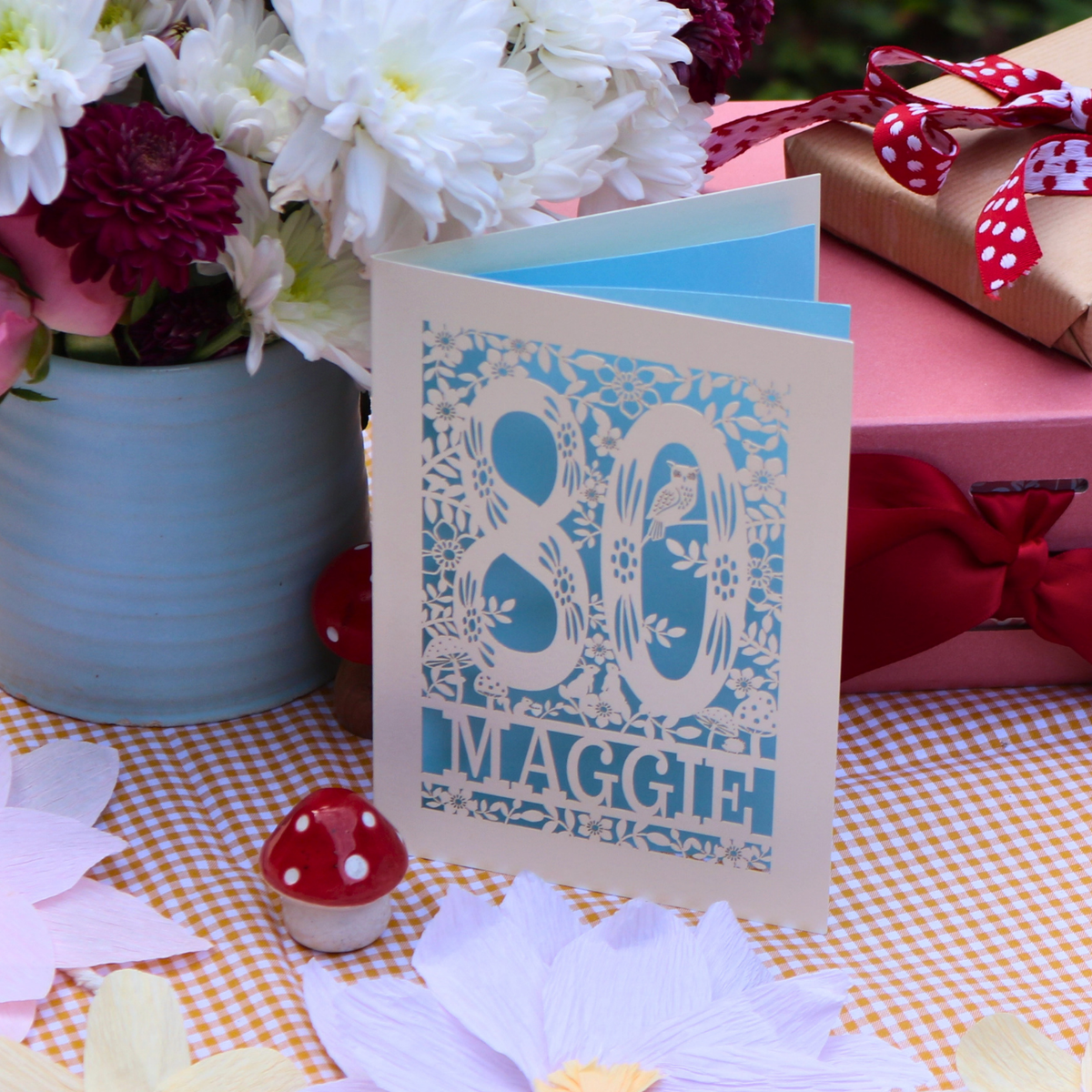A laser cut greeting card with a blue floral pattern and the text '80th Maggie' stands on a checked tablecloth. The card is styled with paper flowers, a small red mushroom figurine, a pink gift box with a red polka-dot ribbon, and a vase of white and burgundy flowers in the background.