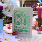 A handmade 80th birthday card with a green and cream floral cut out design, displayed on a checked tablecloth surrounded by flowers, candles, and small mushroom figurines.