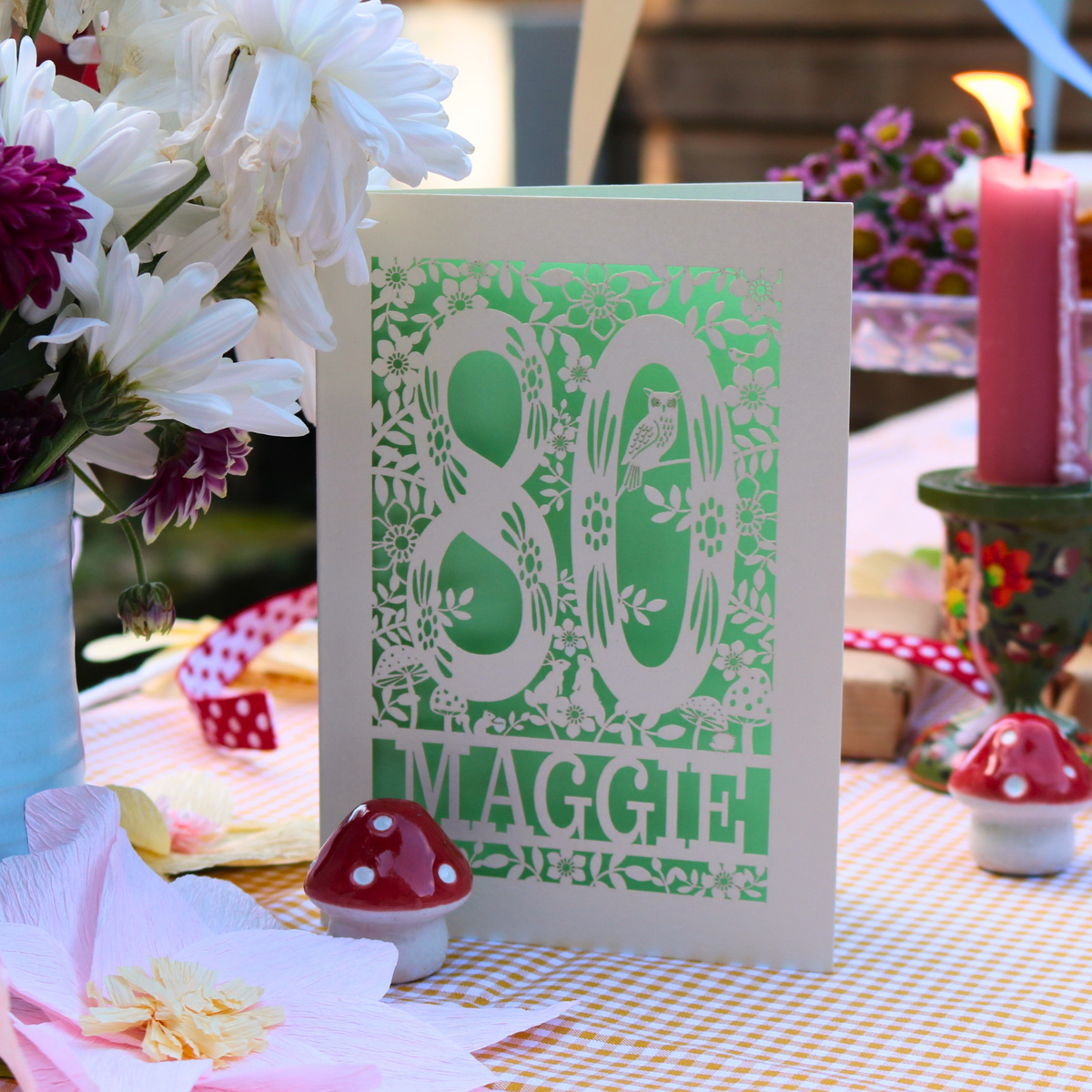 A handmade 80th birthday card with a green and cream floral cut out design, displayed on a checked tablecloth surrounded by flowers, candles, and small mushroom figurines.