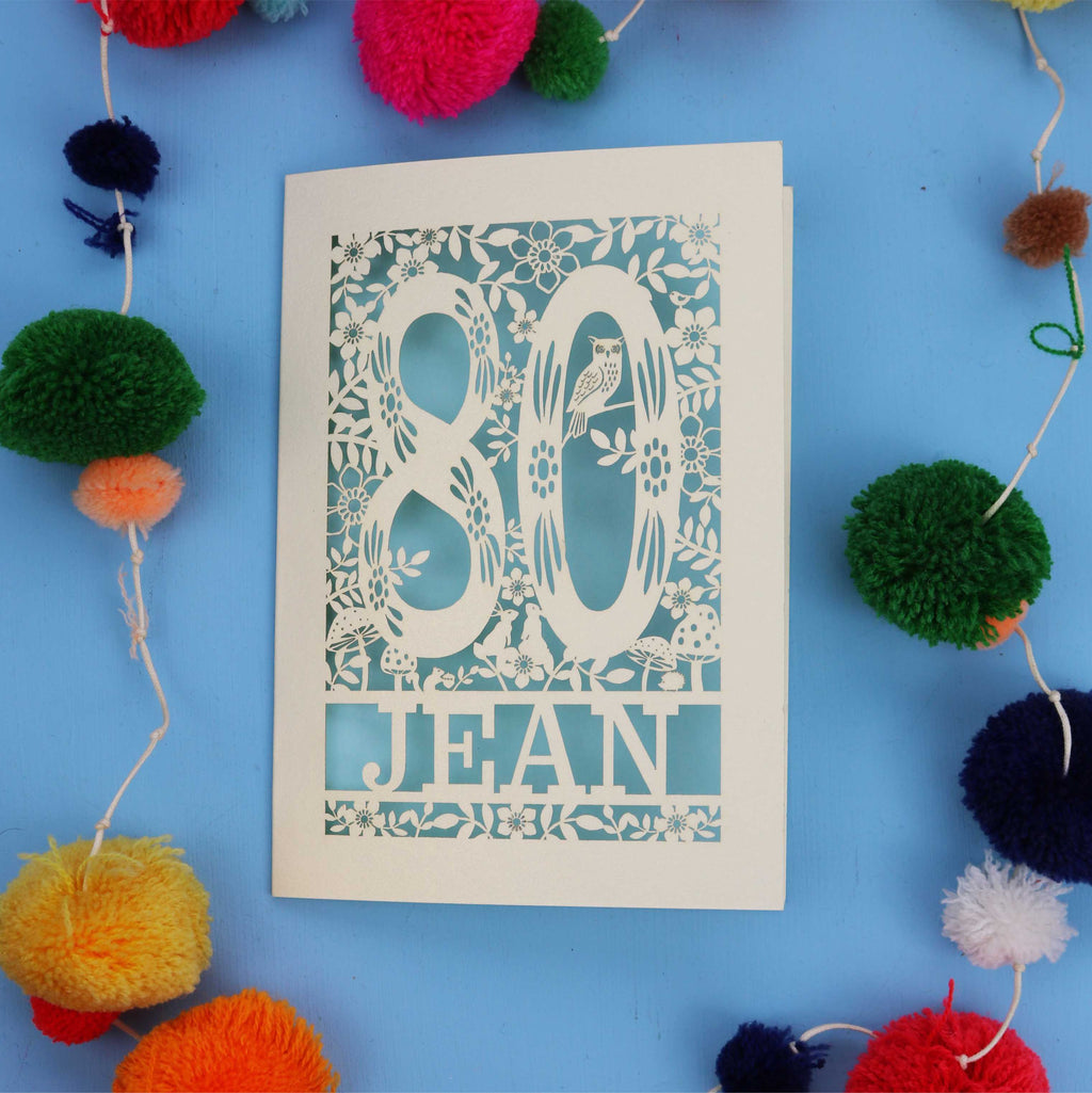 Unique 80th Birthday card with '80 Jean' design on a blue background with colourful pom-poms.