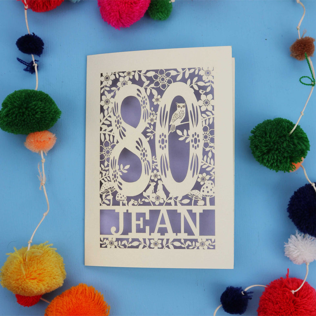 Paper Cut 80th Birthday card with '80 Jean' design on a blue background with colourful pom-poms.