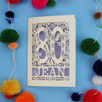 Paper Cut 80th Birthday card with '80 Jean' design on a blue background with colourful pom-poms.