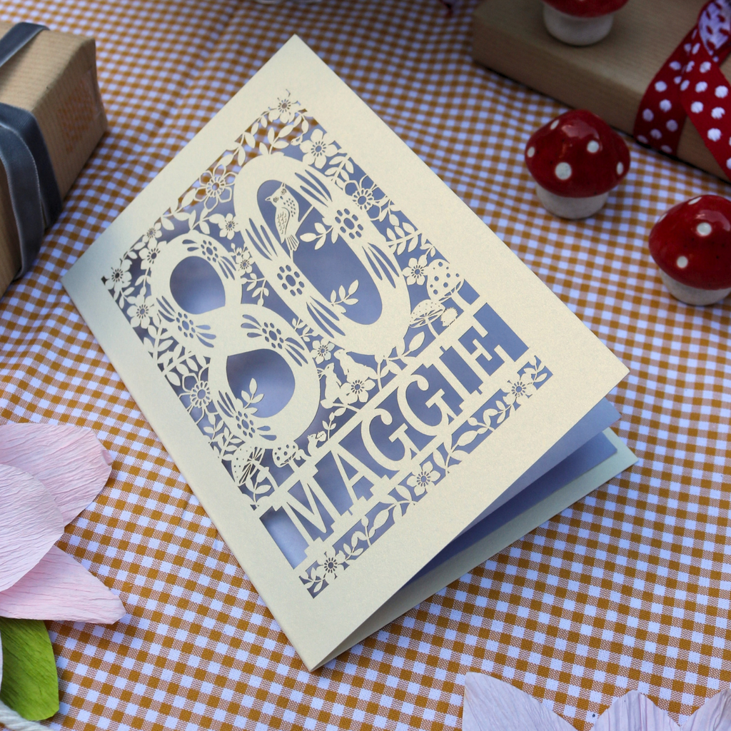 A cream-colored 80th birthday card with intricate laser-cut floral and bird designs around the number 80 and the name 'Maggie' cut out, displayed on a brown and white checkered tablecloth with small decorative props.