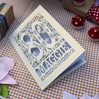 A cream-colored 80th birthday card with intricate laser-cut floral and bird designs around the number 80 and the name 'Maggie' cut out, displayed on a brown and white checkered tablecloth with small decorative props.