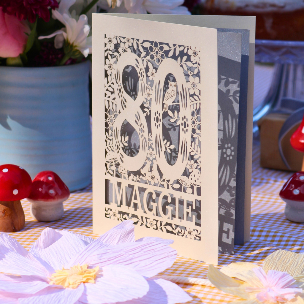 A personalised 80th birthday card with intricate laser-cut floral and woodland patterns and the name 'Maggie' at the bottom, displayed on a checked tablecloth with decorative flowers and small red mushrooms.