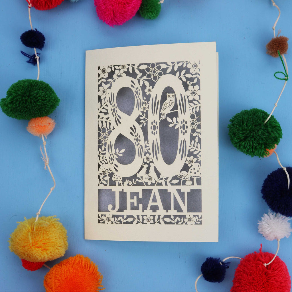 Decorative 80th Birthday card with '80 Jean' design on a blue background with colourful pom-poms.