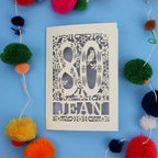 Decorative 80th Birthday card with '80 Jean' design on a blue background with colourful pom-poms.