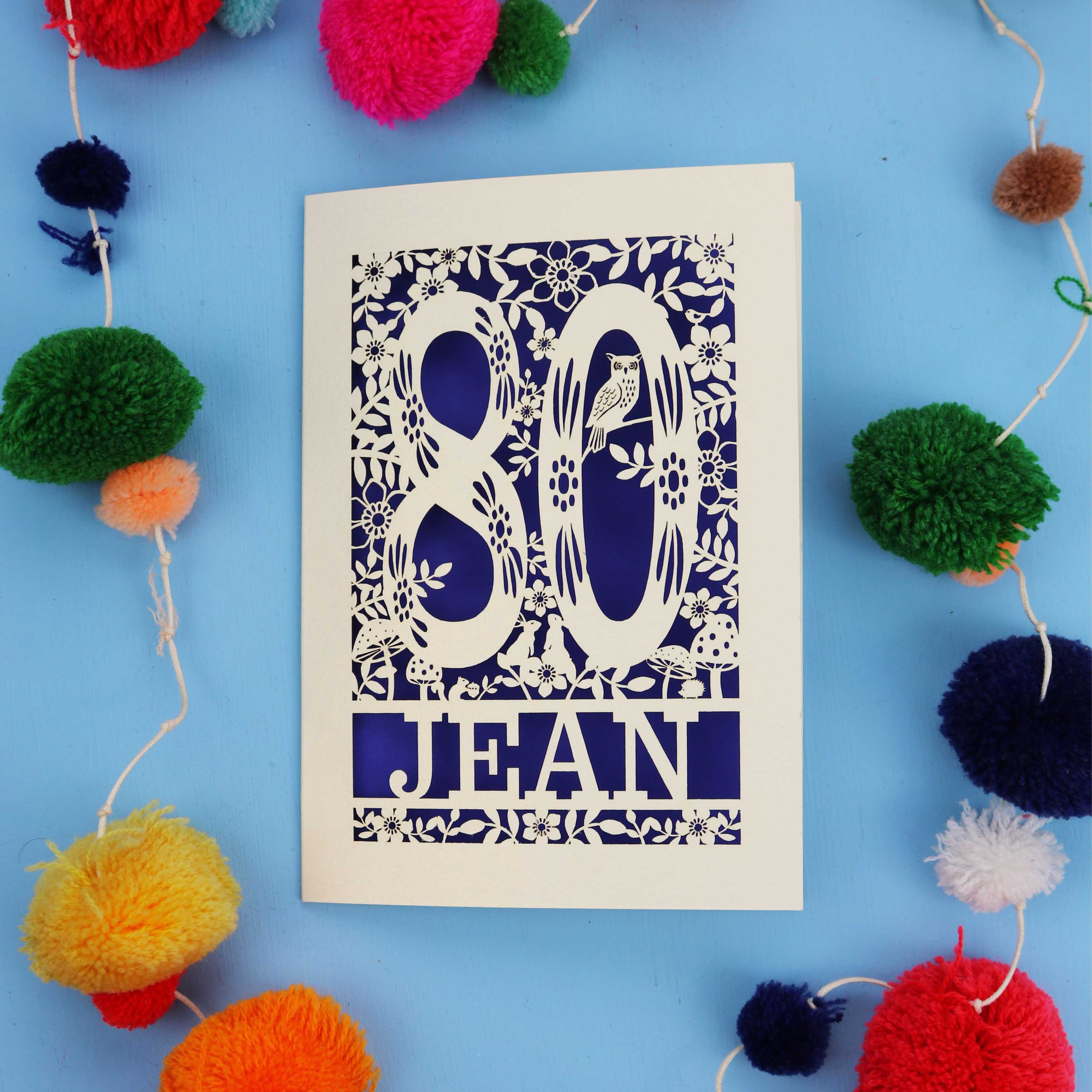 Woodland 80th Birthday card with '80 Jean' design on a blue background with colourful pom-poms.